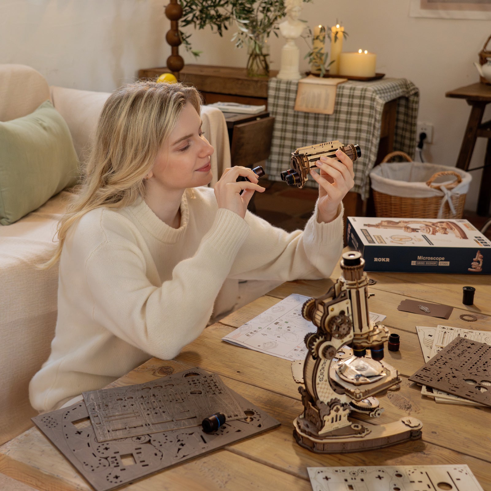 Woman in a cream sweater closely inspecting the ROKR wooden microscope model while seated at a cozy home workspace