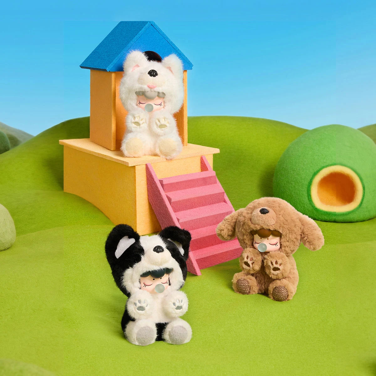 Three plush figures in black-and-white, cream, and brown dog costumes posed around a yellow felt house with a blue roof and pink stairs on green felt.