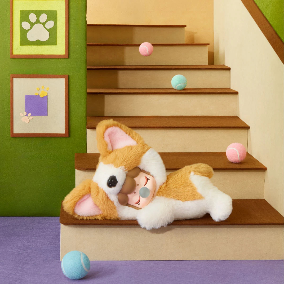 Plush corgi toy with a doll face and bubble lying on miniature wooden stairs, surrounded by pink and blue tennis balls, paw print wall art and green walls.