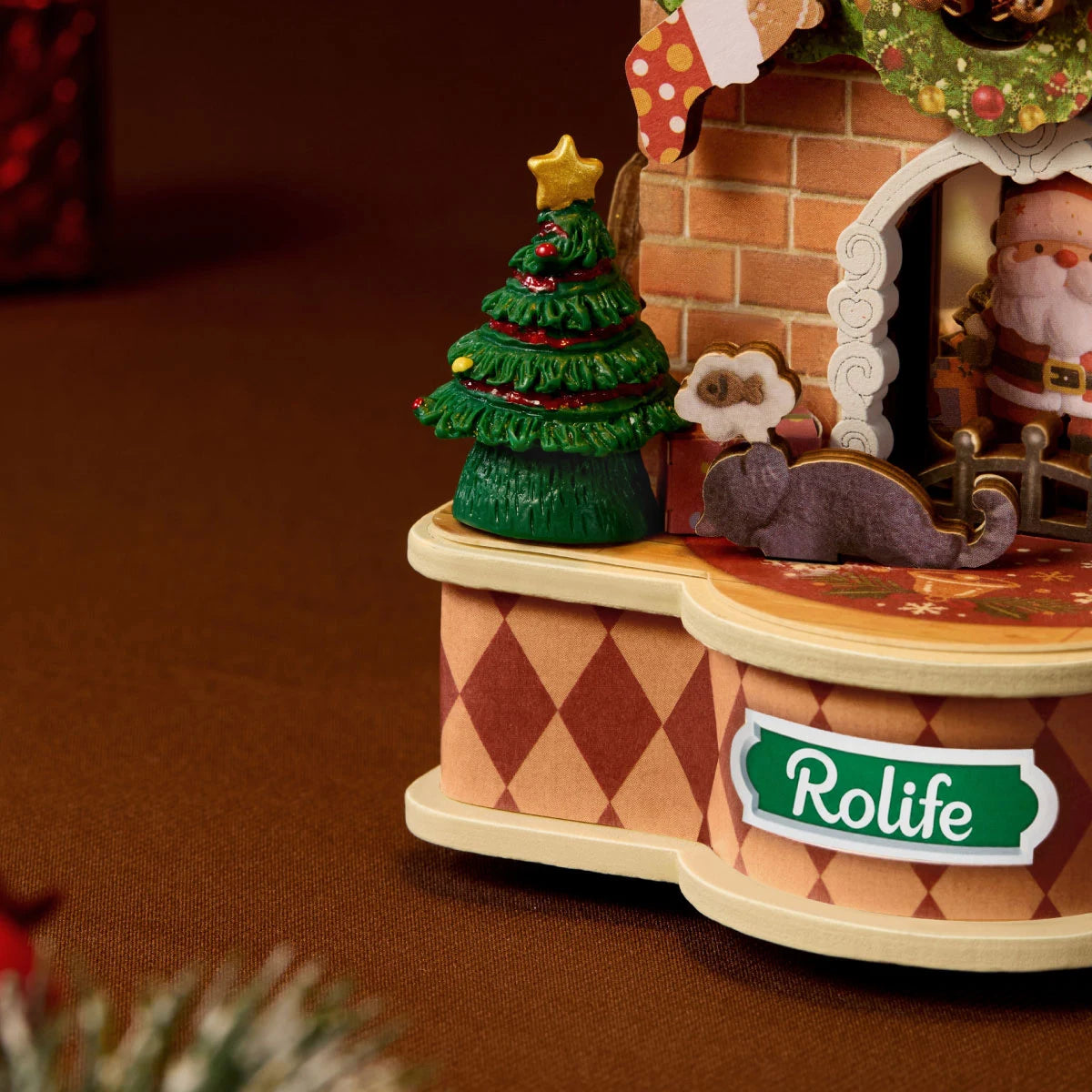 Close-up of the Rolife model base with green Rolife sign, diamond-patterned sides, miniature Christmas tree with star topper, grey cat and Santa figure inside the fireplace.