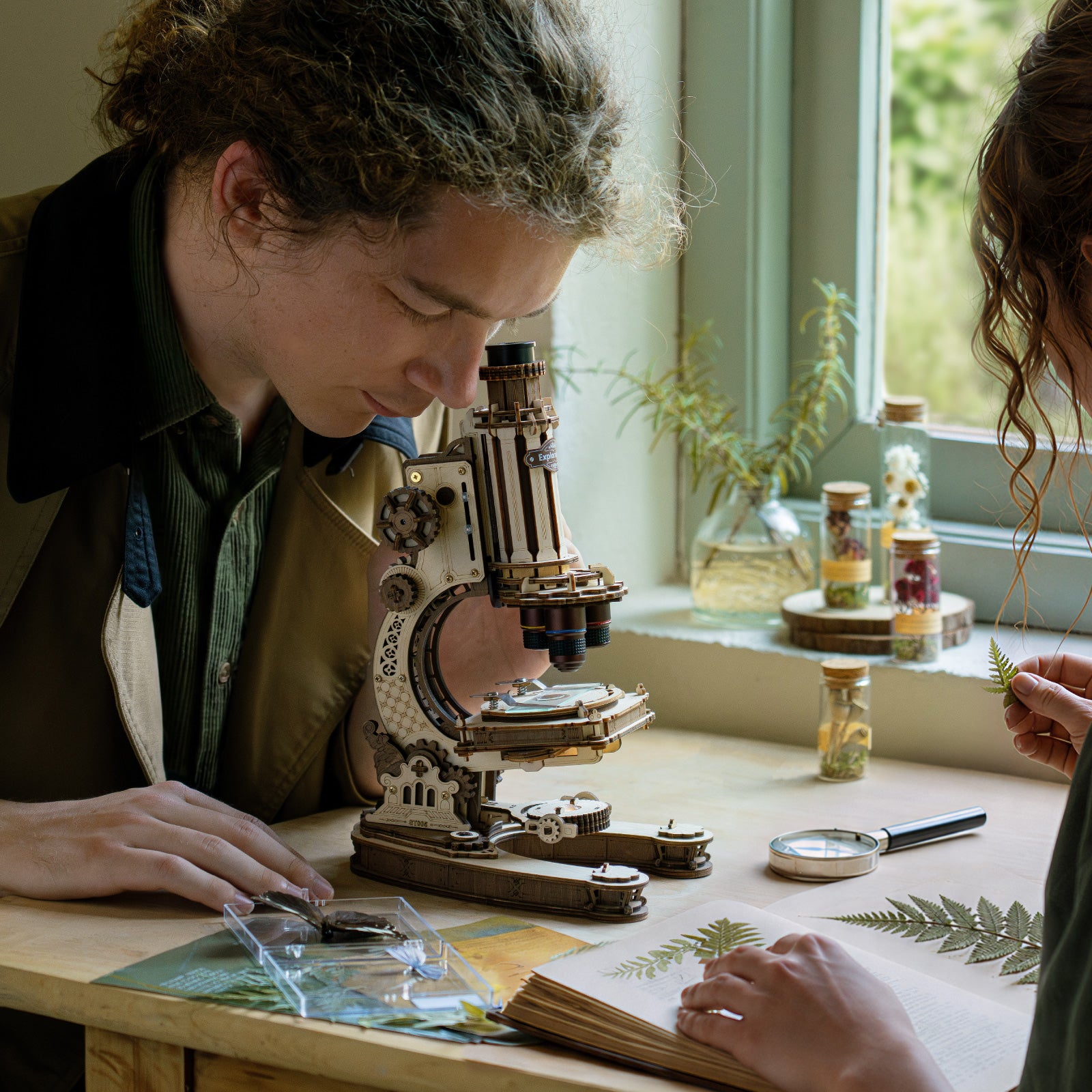 Two people examining botanical specimens on slides under the ROKR wooden microscope at a sunlit window ledge