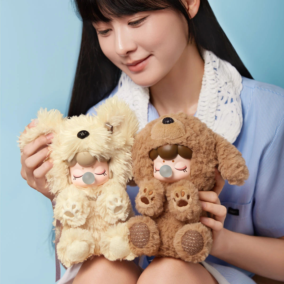 Person holding two plush figures in cream and brown dog costumes with closed eyes and bubble accessories against a light blue background.