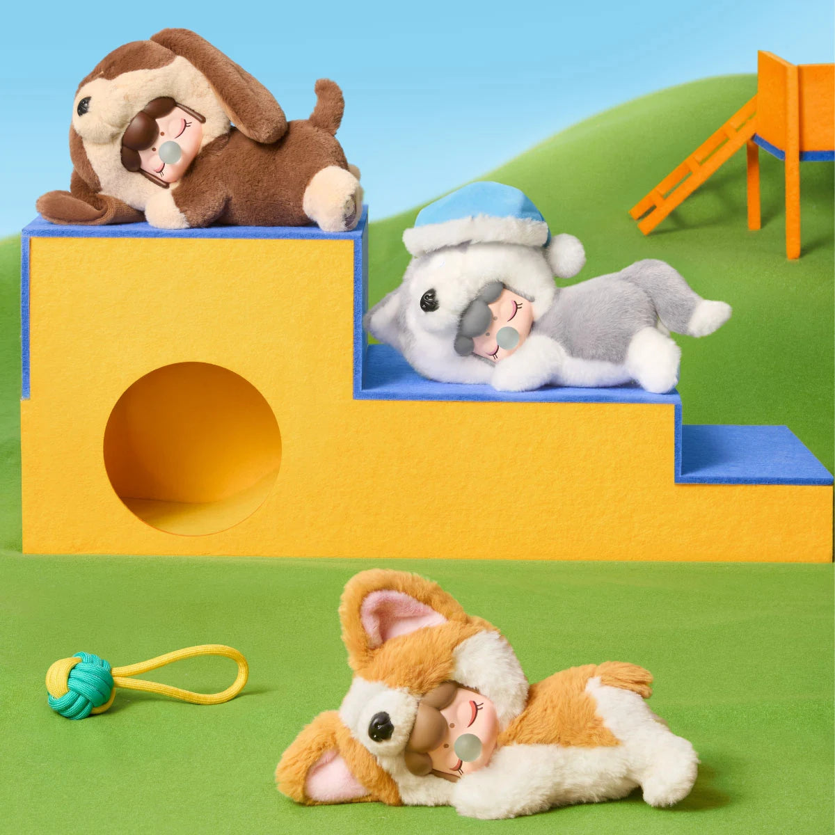 Three plush figures in brown, grey, and orange dog costumes resting on yellow and blue felt blocks on a green felt surface with a rope toy.