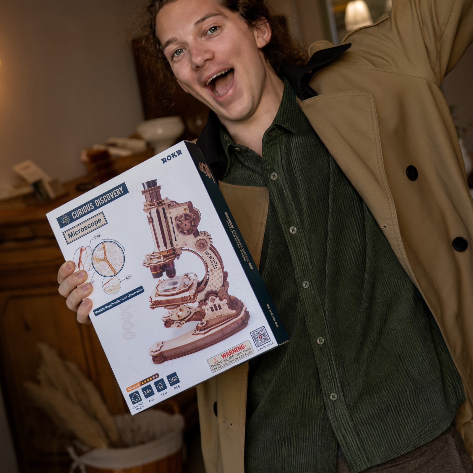 Excited young man holding a ROKR wooden microscope kit box in a warm indoor setting, ready to build the model