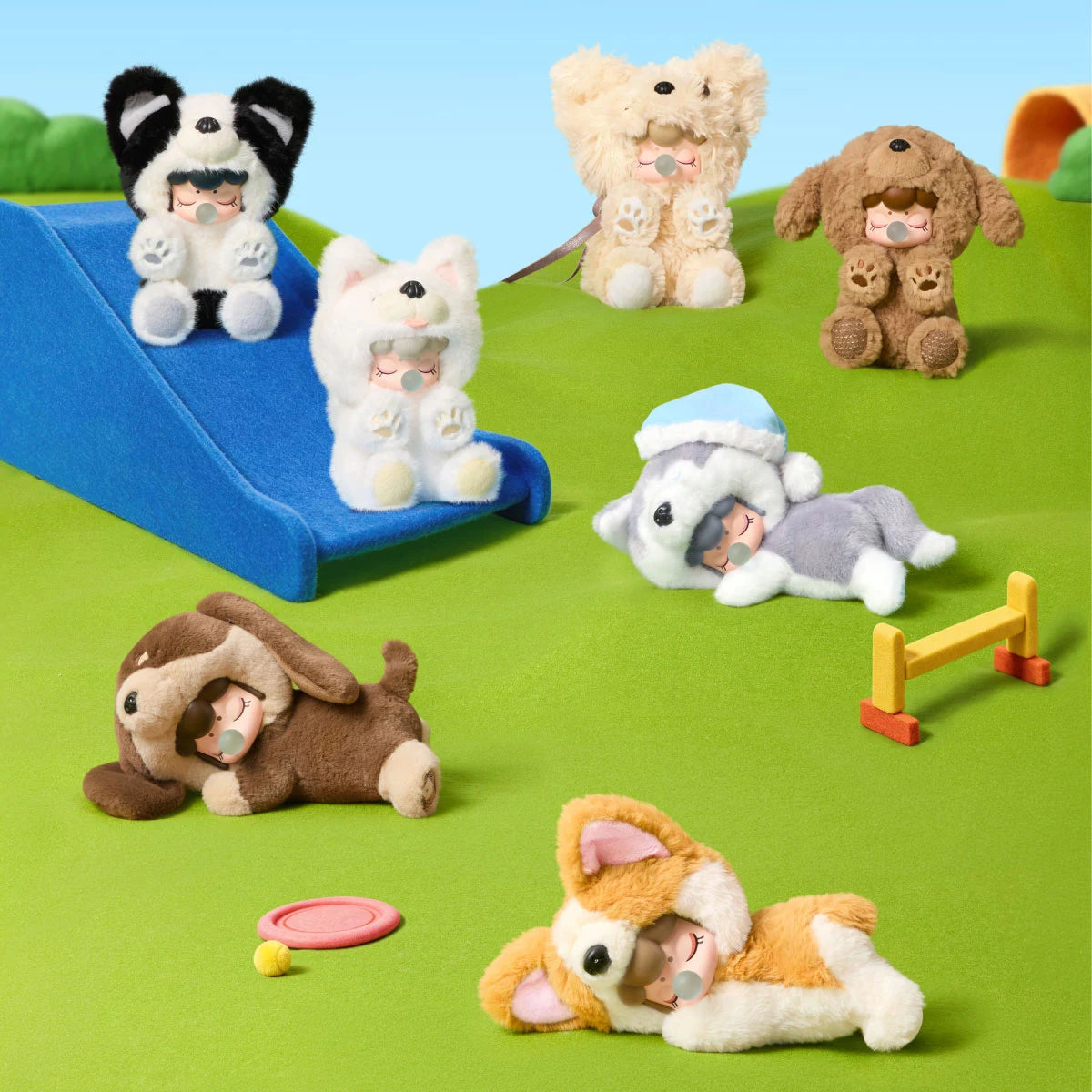 Six plush figures in dog costumes with closed eyes and bubble accessories posed on a green felt playground with a blue slide, seesaw, and ball.