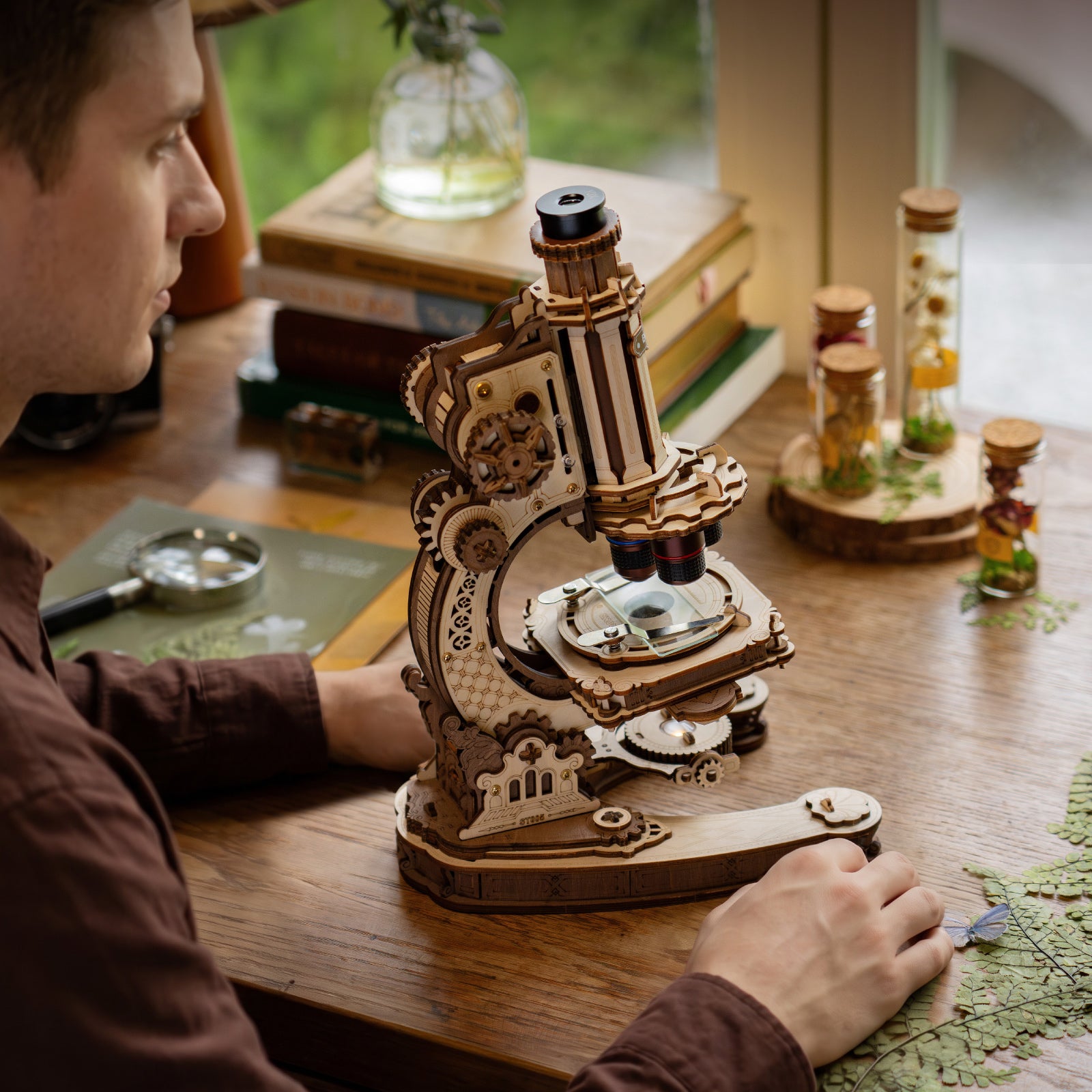 Person studying at a wooden desk with the ROKR wooden microscope in the foreground surrounded by books, plants and glass bottles