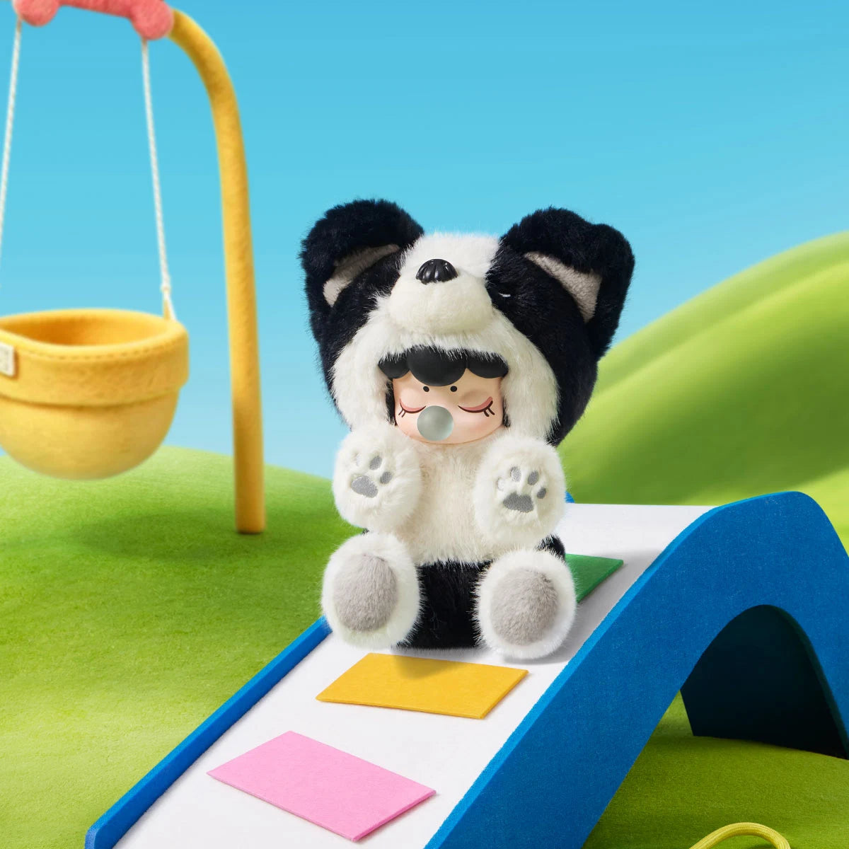 Plush figure in a black-and-white dog costume with closed eyes and a bubble accessory sitting on a blue slide with a yellow swing in the background.