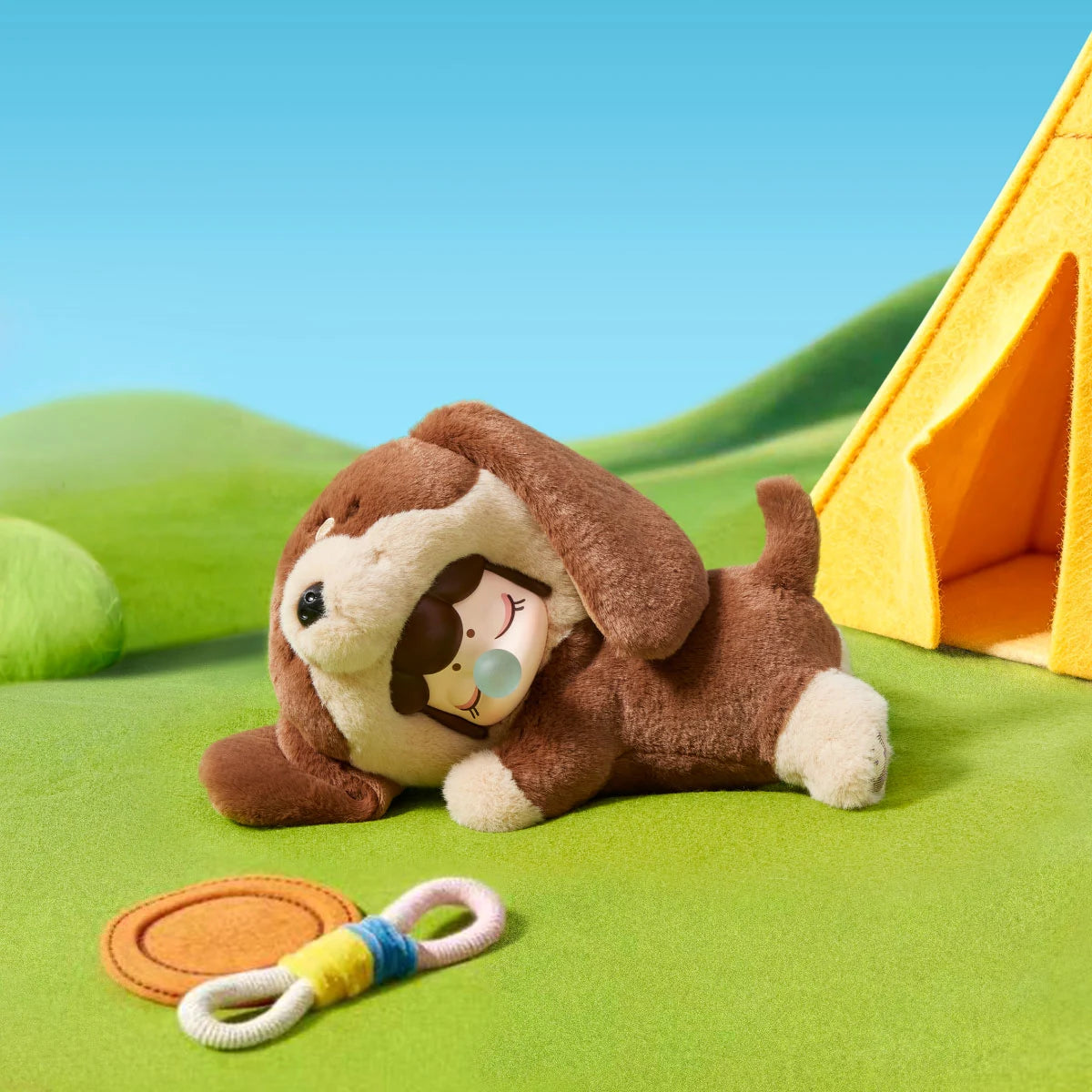 Brown and cream plush puppy toy with a doll face resting on green felt grass beside a yellow tent, with a rope toy and orange disc nearby.
