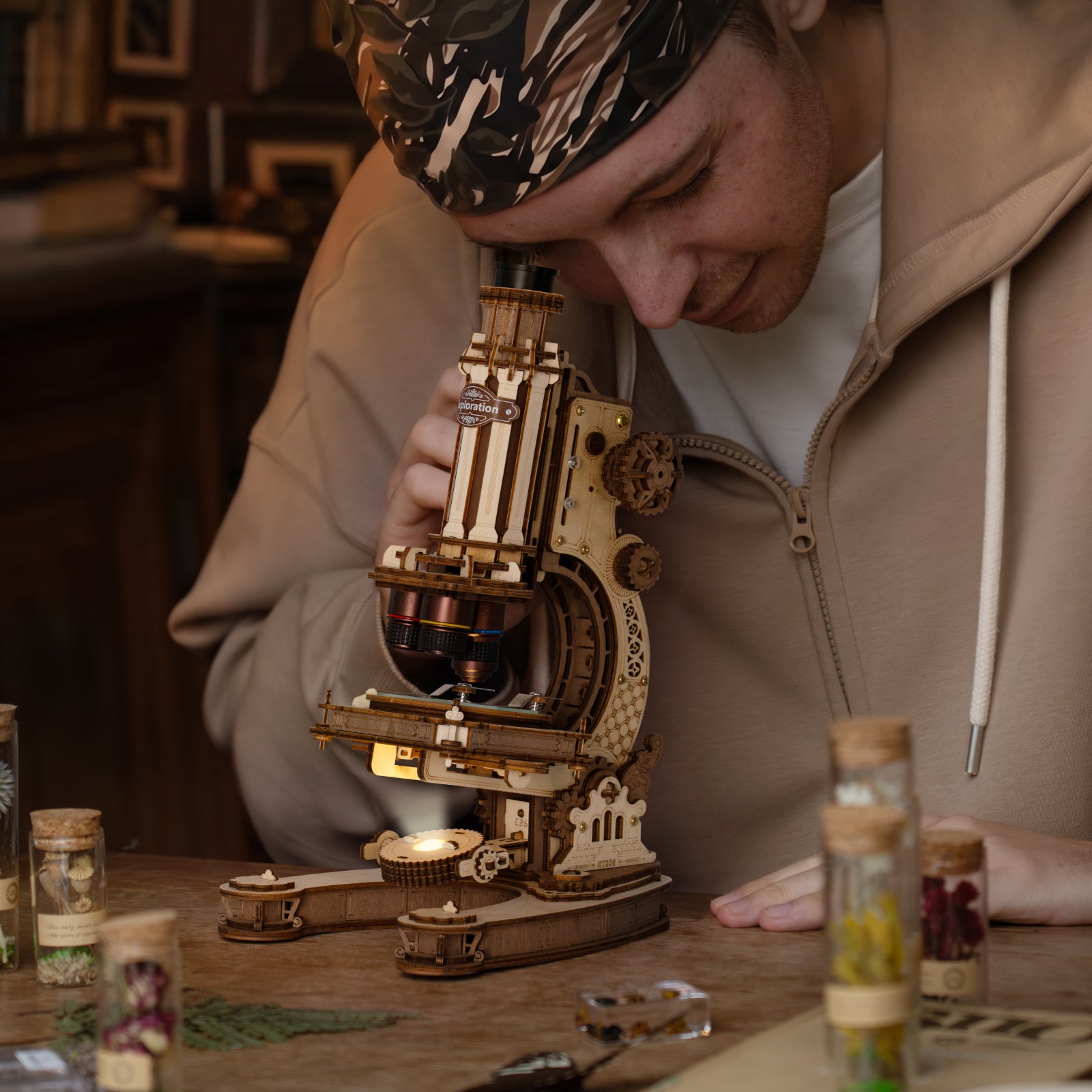 Person in a beige coat leaning over the ROKR wooden microscope on a vintage desk with small glass vials and warm ambient light