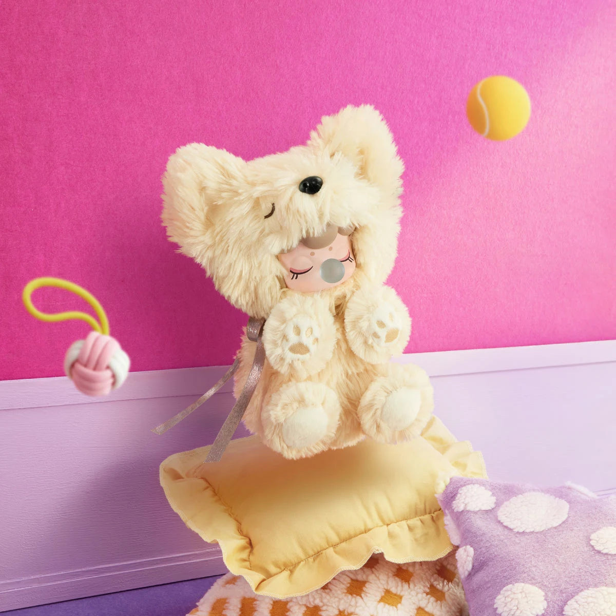 Cream plush dog toy with a doll face sitting on a yellow cushion against a pink wall, with a yellow tennis ball and pink rope toy.