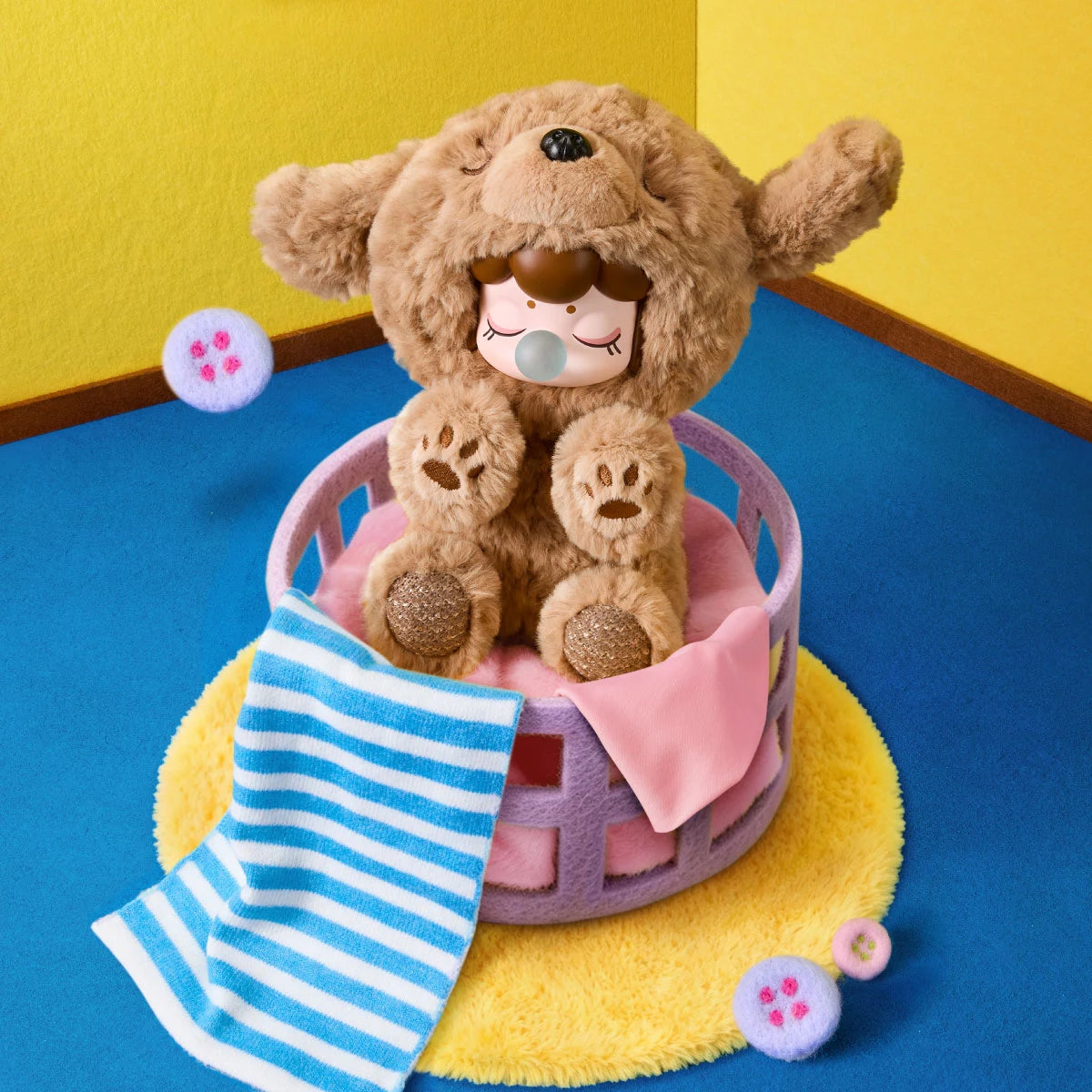 Plush brown puppy-hooded figure with closed eyes and blue bubble, seated in a lavender basket on a yellow rug, with striped towel and pink cloth.