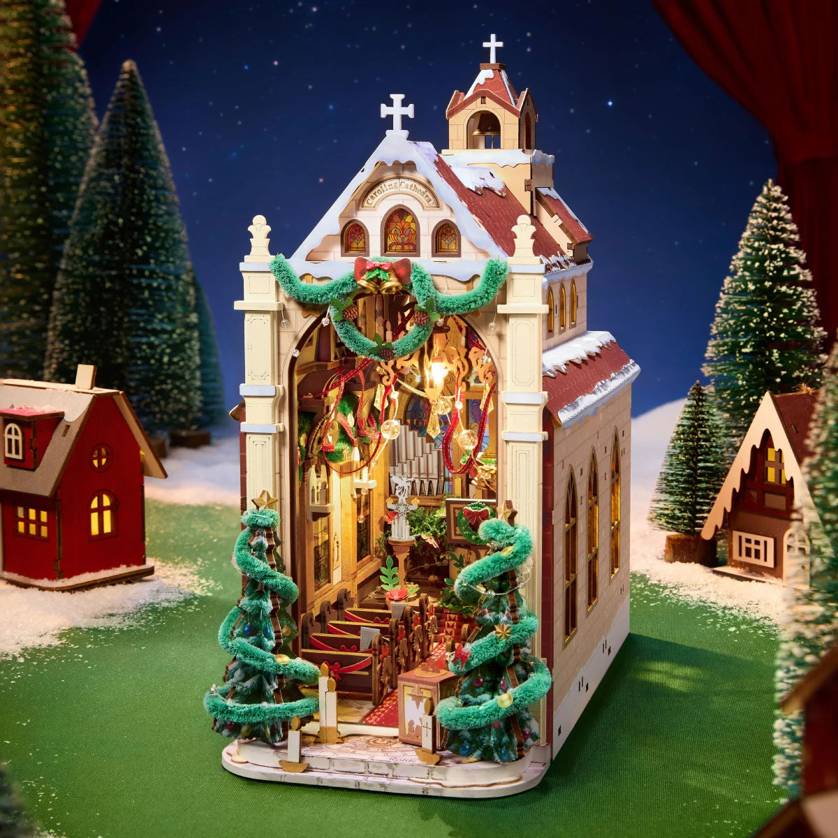 Rolife miniature Christmas cathedral with open front revealing a lit interior, flanked by decorated trees on a snowy green base with small houses.