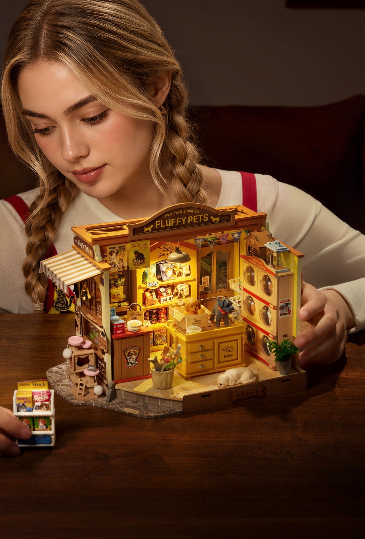 Woman assembling a warmly lit miniature corner shop with shelves and a yellow awning