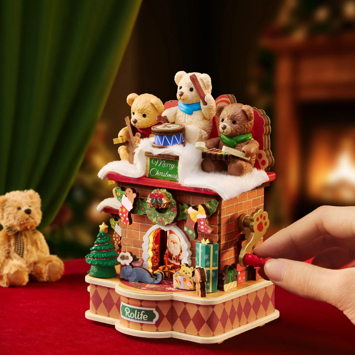 Hand turning the red crank handle on the side of the Rolife Christmas fireplace music box, with teddy bears on top and a plush bear beside it on red fabric.