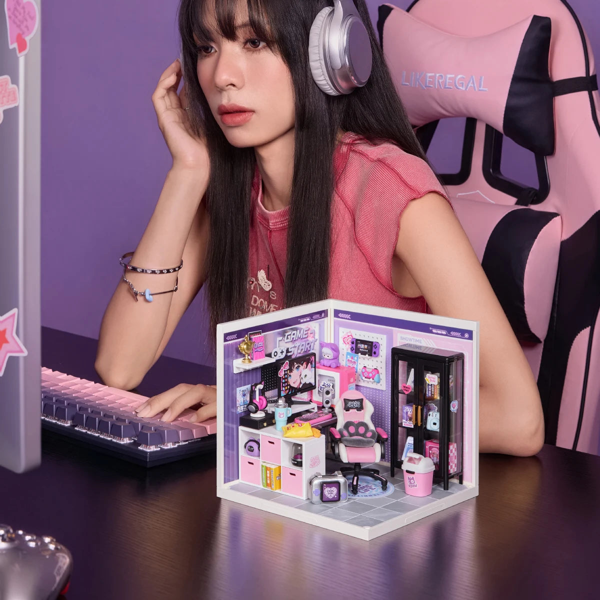 Miniature gaming room diorama placed on a desk beside a pink keyboard, with a person in a pink gaming chair in the background.