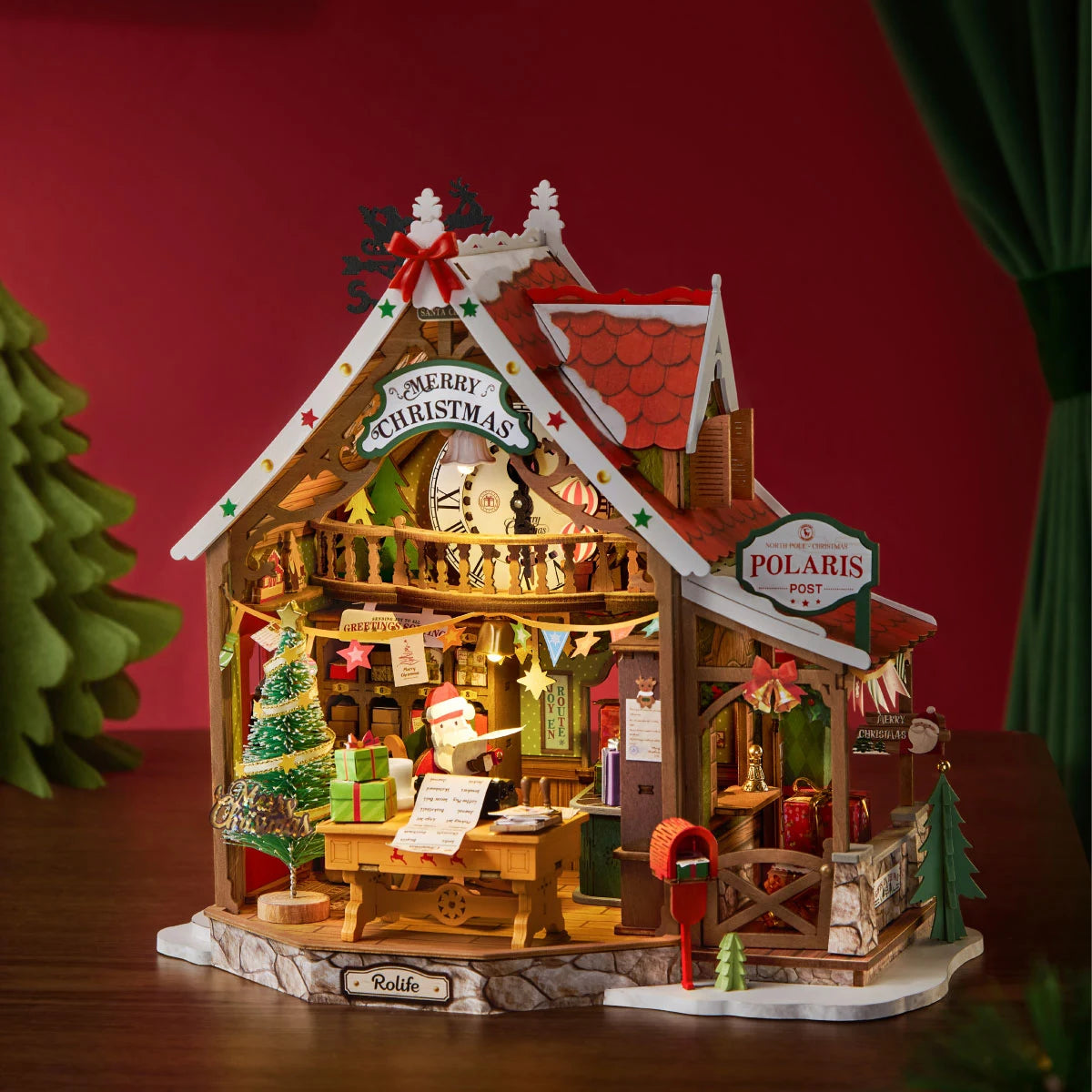 Rolife wooden miniature Christmas post office with red roof, Santa figure at desk, decorated tree, red mailbox and Polaris Post sign, lit warmly against a red backdrop.
