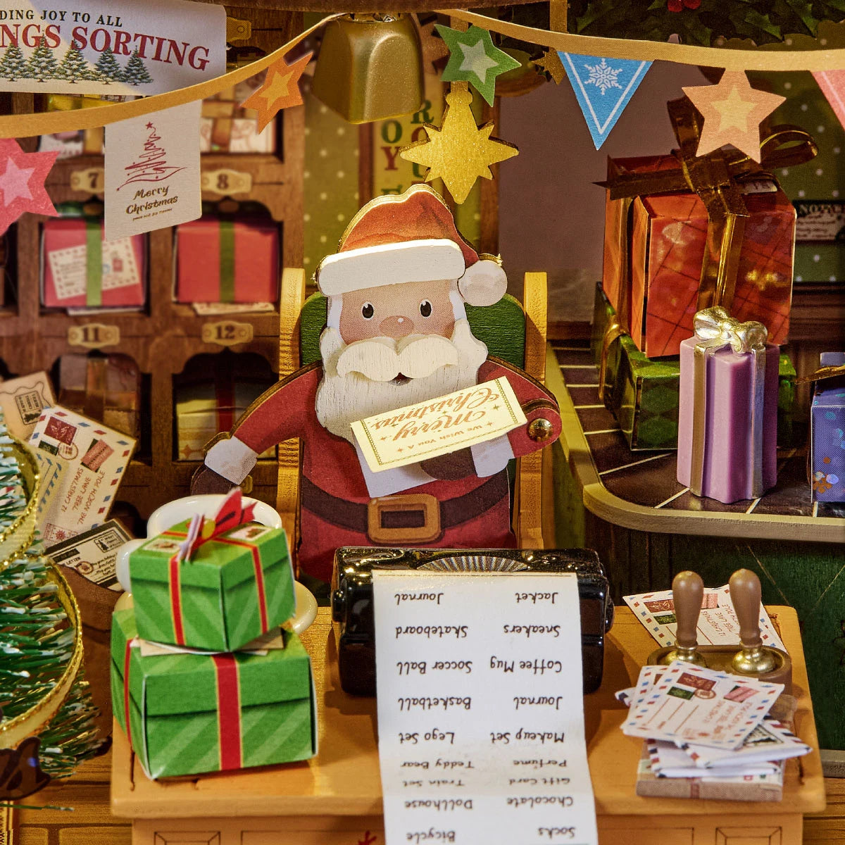 Close-up of a wooden Santa figure holding a Merry Christmas card at a desk with a list, green wrapped gifts, letters and festive bunting inside the miniature post office.