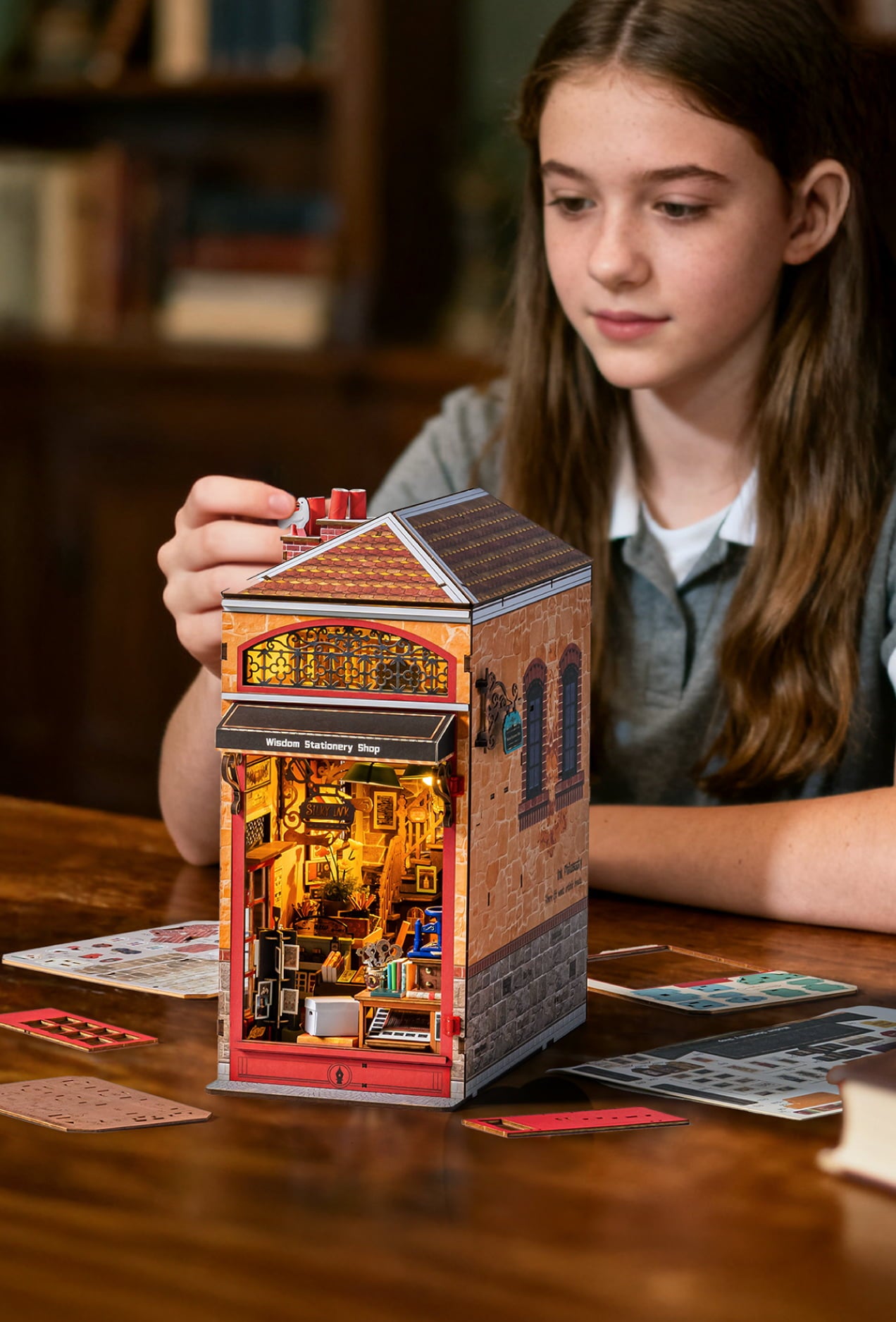 Young woman adjusting the roof of a tall illuminated miniature shop on a wooden table