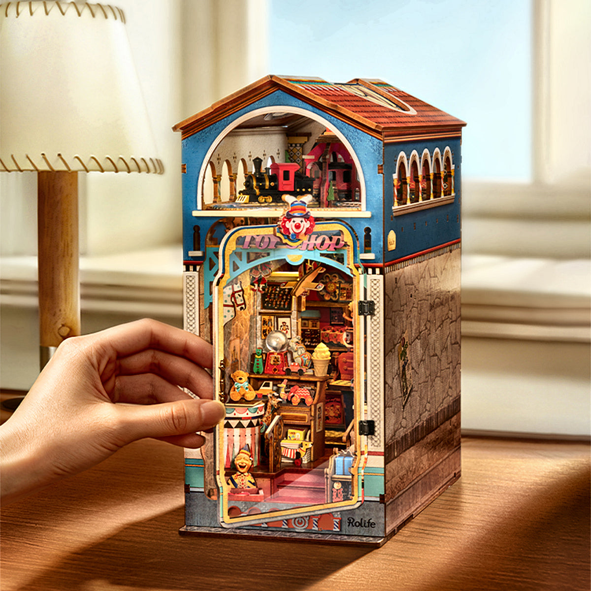 Hand adjusting the colorful Treasure Toy Shop book nook beside a cream table lamp