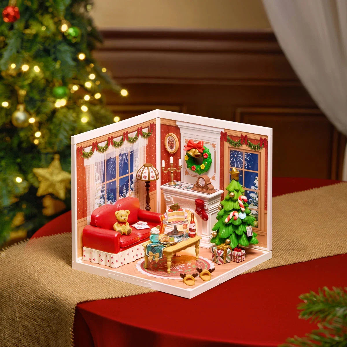 Rolife miniature Christmas living room diorama on a red table runner, showing red sofa, fireplace, Christmas tree and lit windows.